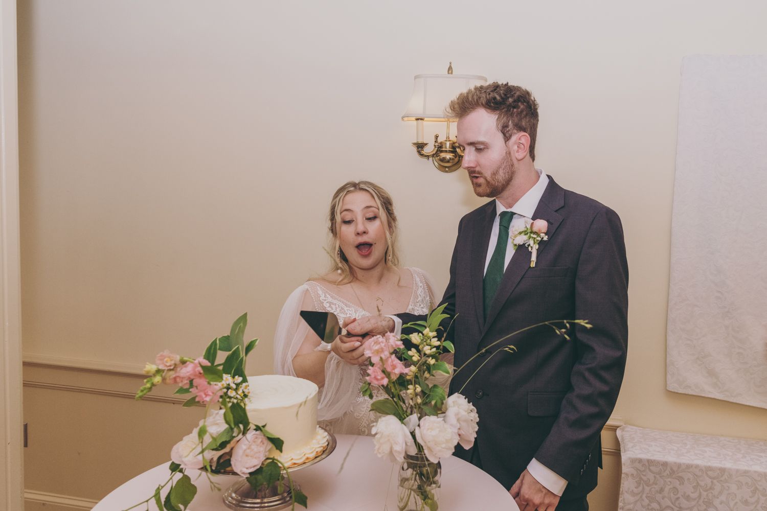 Bride and groom laugh joyfully while cutting their floral-decorated wedding cake at their indoor reception.
