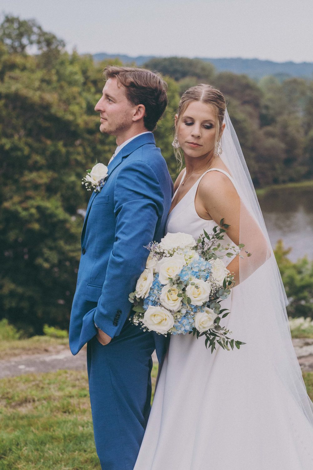 Bride and groom stand back-to-back outdoors, she holds white and blue bouquet, he wears blue suit, lush greenery behind them.