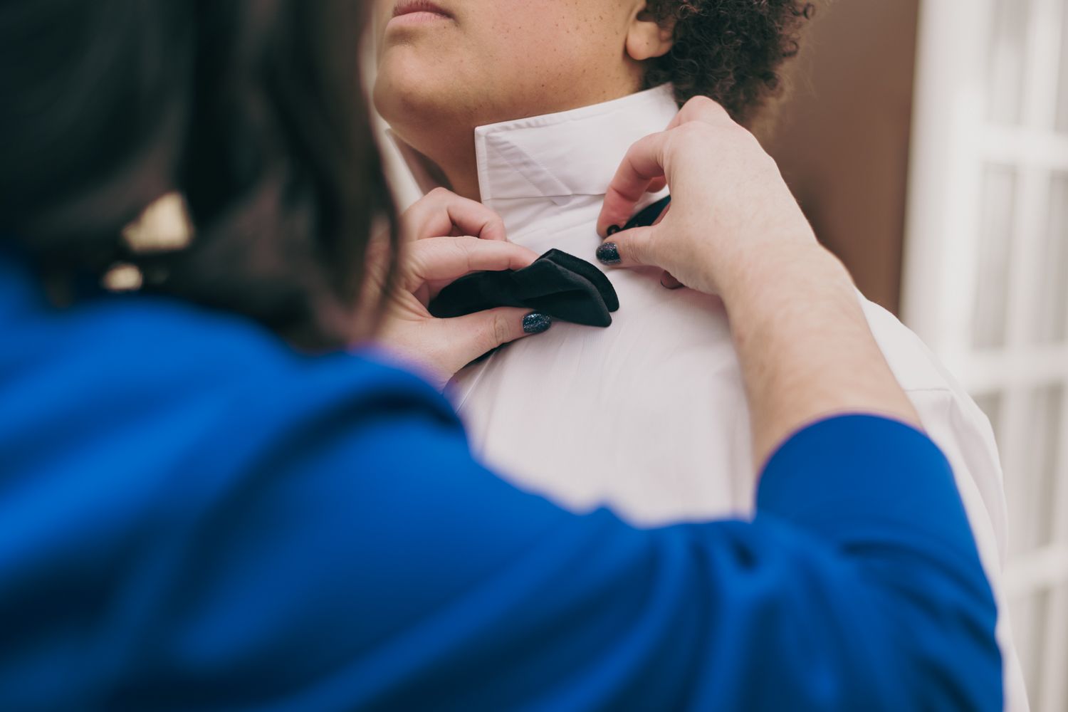 Woman in blue dress fastening a black bow tie on a groom in a white dress shirt.