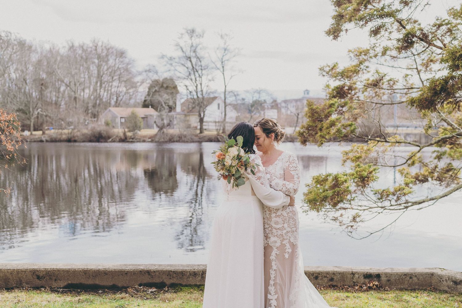 Two brides in elegant wedding gowns embrace near a serene lake surrounded by lush trees.