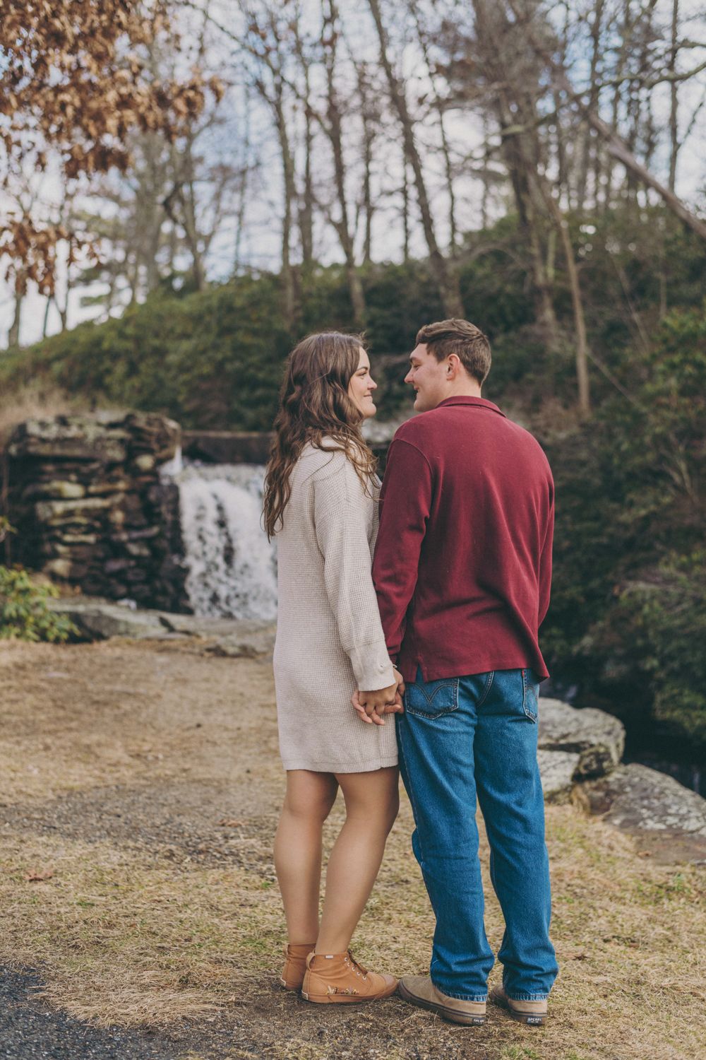 A couple holds hands and gazes at each other near a waterfall surrounded by trees in autumn.