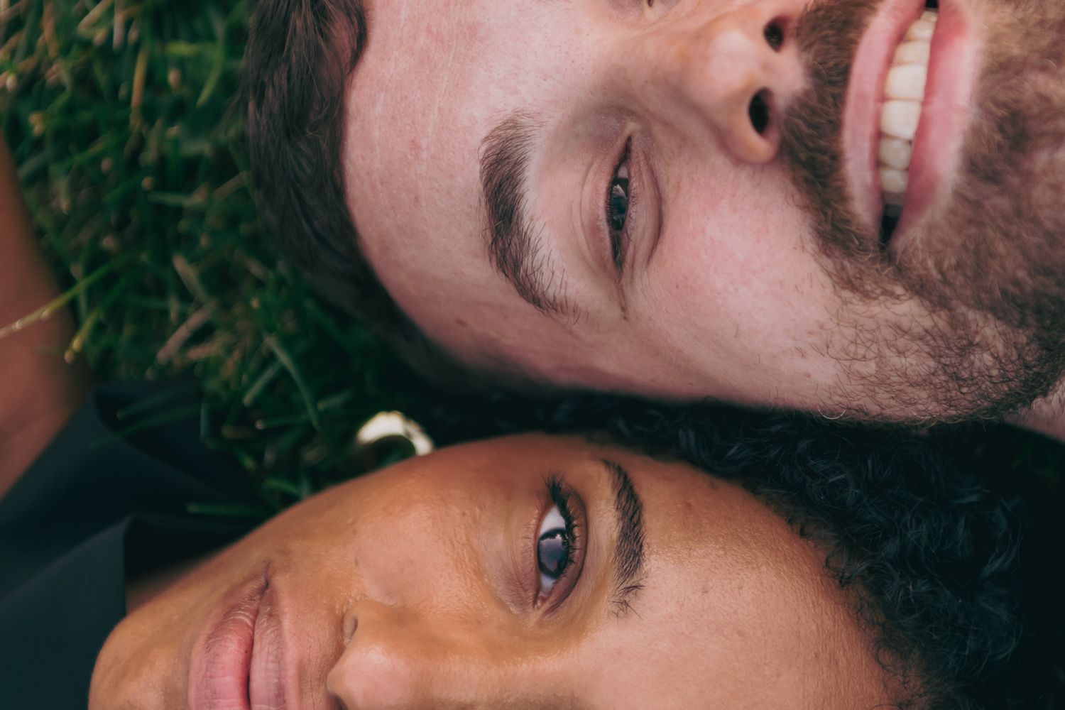 Close-up portrait of an interracial couple lying head to head on green grass, smiling warmly.