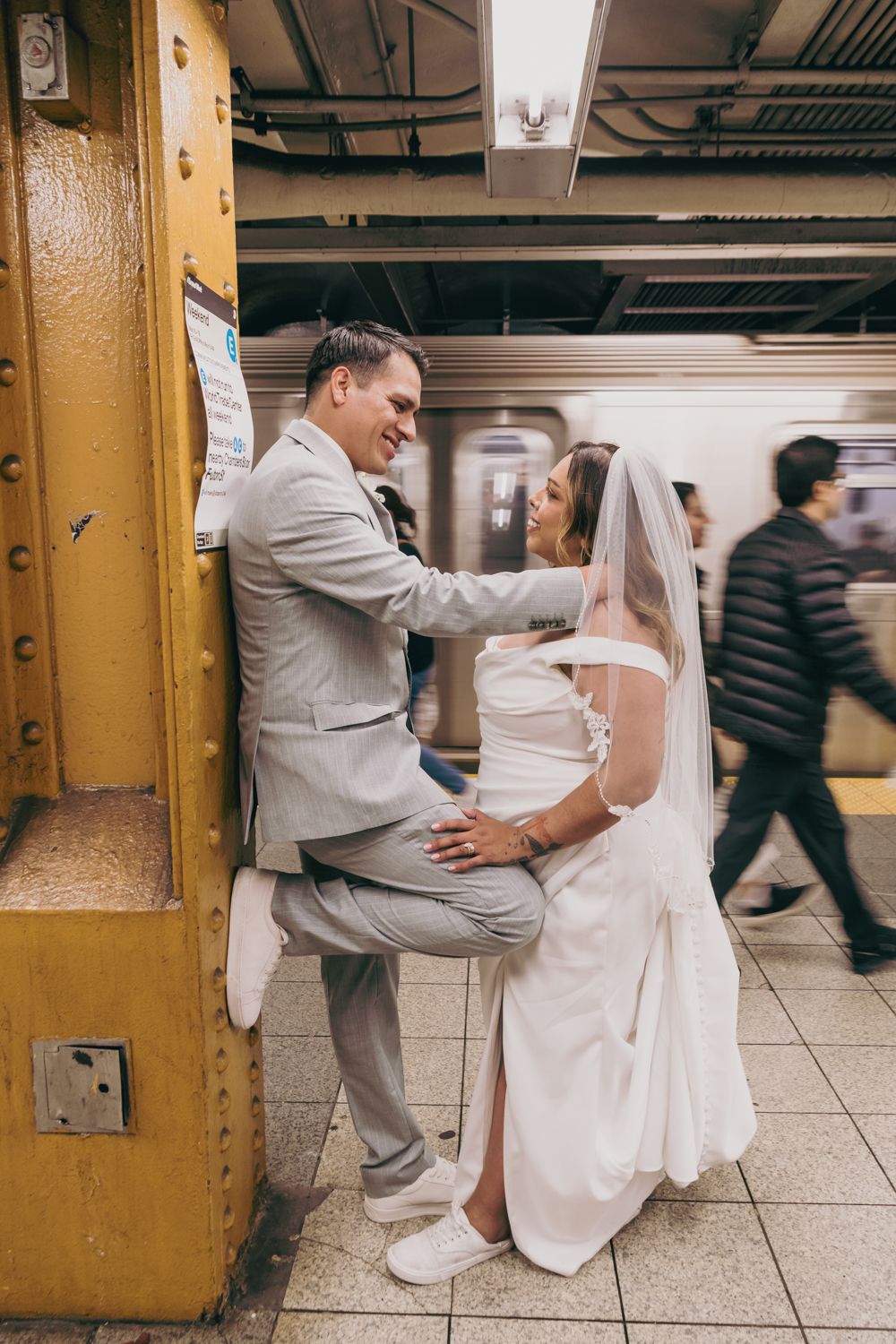 Smiling bride and groom in wedding attire pose playfully against a yellow subway station pillar in NYC.
