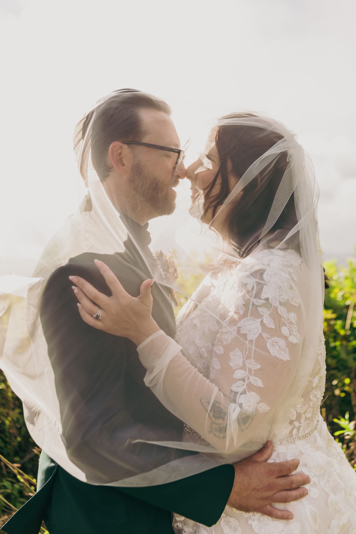 Bride and groom share a romantic moment under a flowing veil, embracing outdoors with soft backlit sunlight.