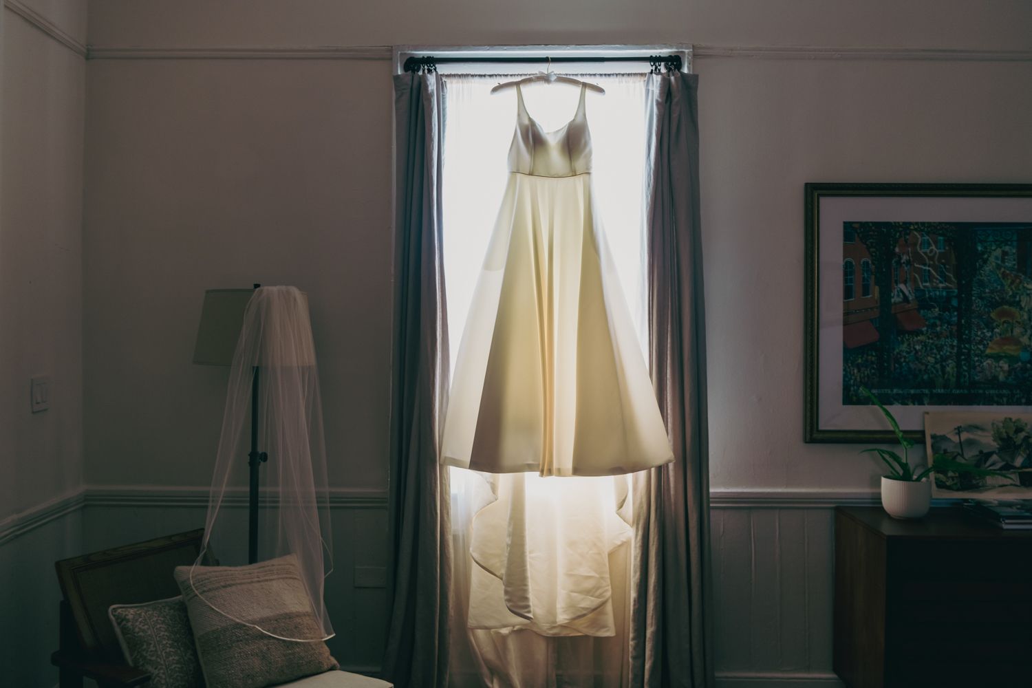Elegant ivory wedding gown hanging in window between gray curtains in softly lit room.