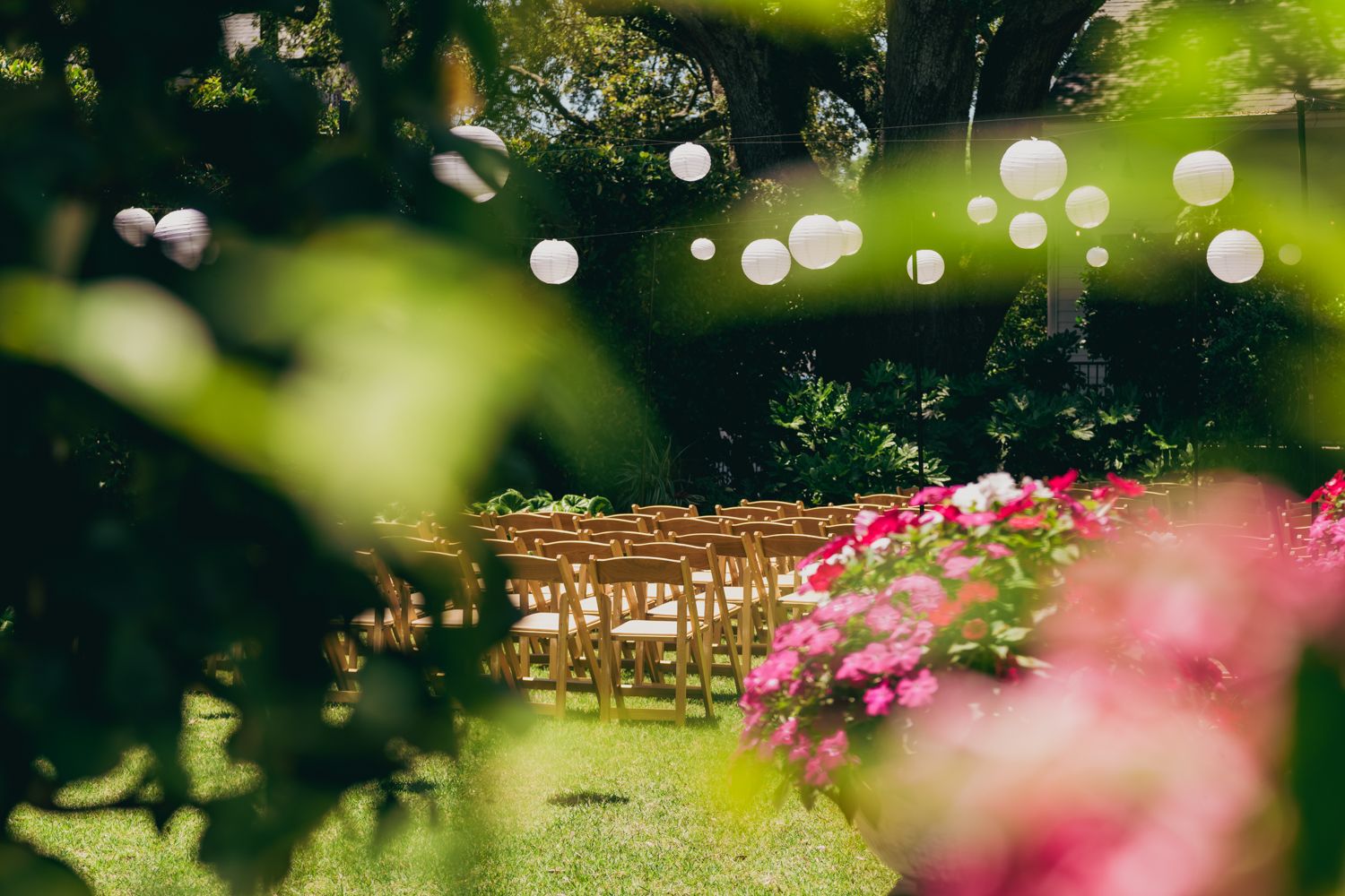 Outdoor wedding ceremony setup with wooden chairs, pink flowers, and white paper lantern decorations.