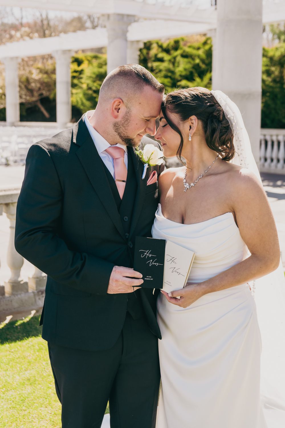 Bride and groom touching foreheads tenderly while holding vow books near white columns on wedding day.