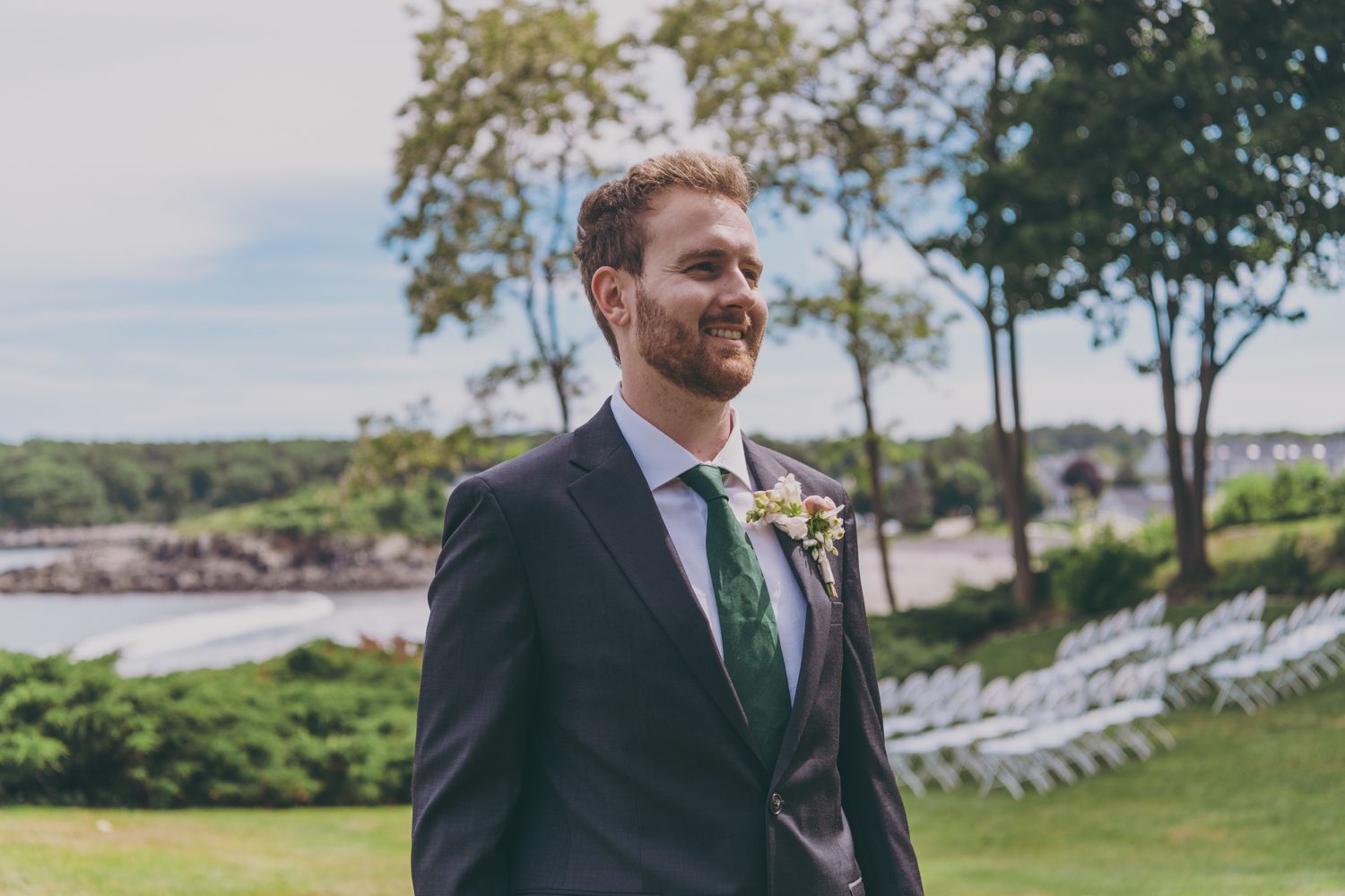 Handsome groom in dark suit with floral boutonniere smiling outdoors at a waterfront wedding venue.