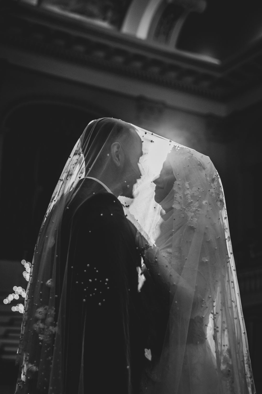 Black and white photo of bride and groom sharing a kiss under a flowing veil in a church.
