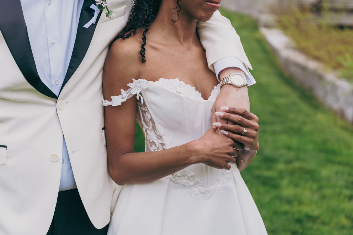 Groom embraces bride in off-shoulder lace wedding gown during romantic outdoor ceremony.