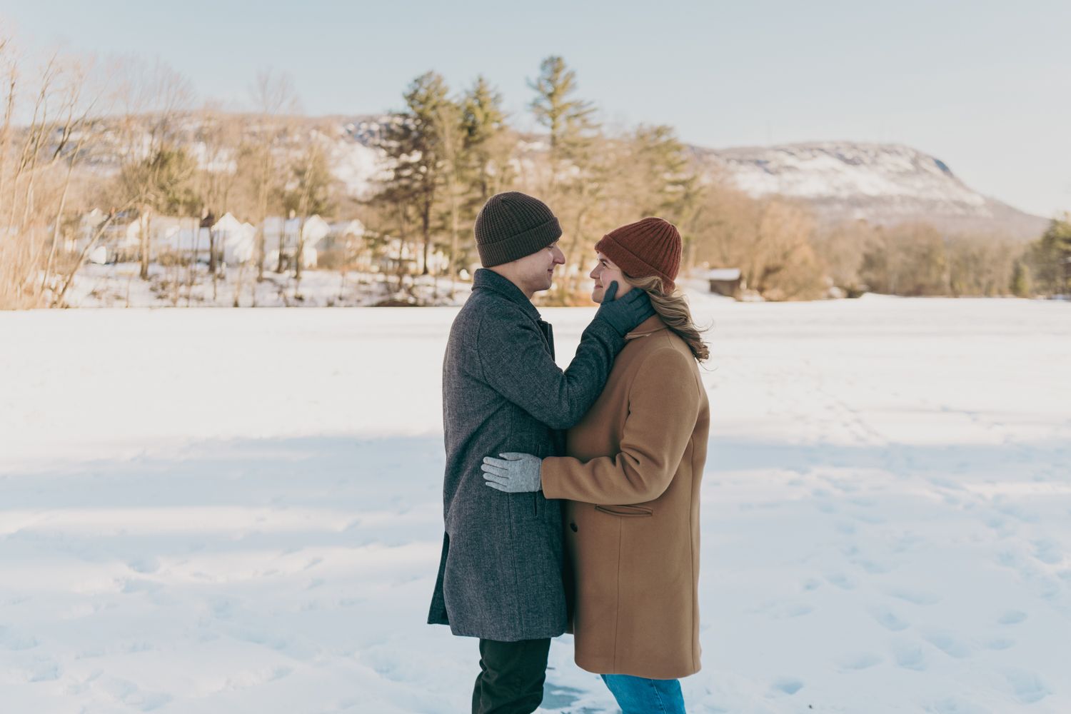 Couple in winter coats sharing a tender moment in a snow-covered field with mountain backdrop.