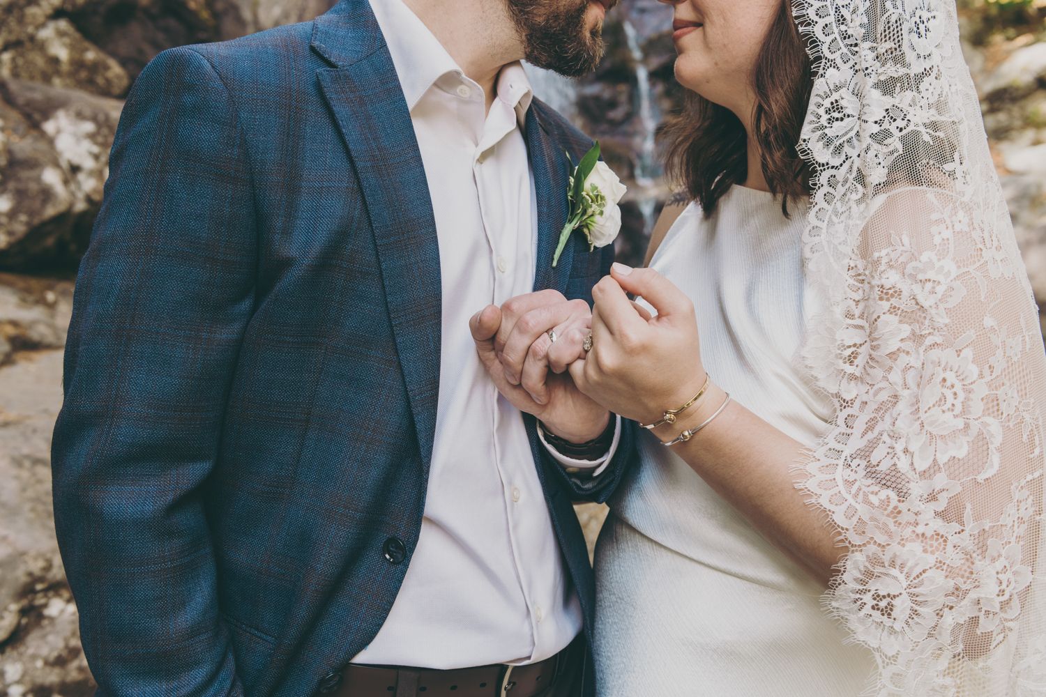 Bride and groom hold hands near a waterfall, showcasing lace veil and boutonniere.