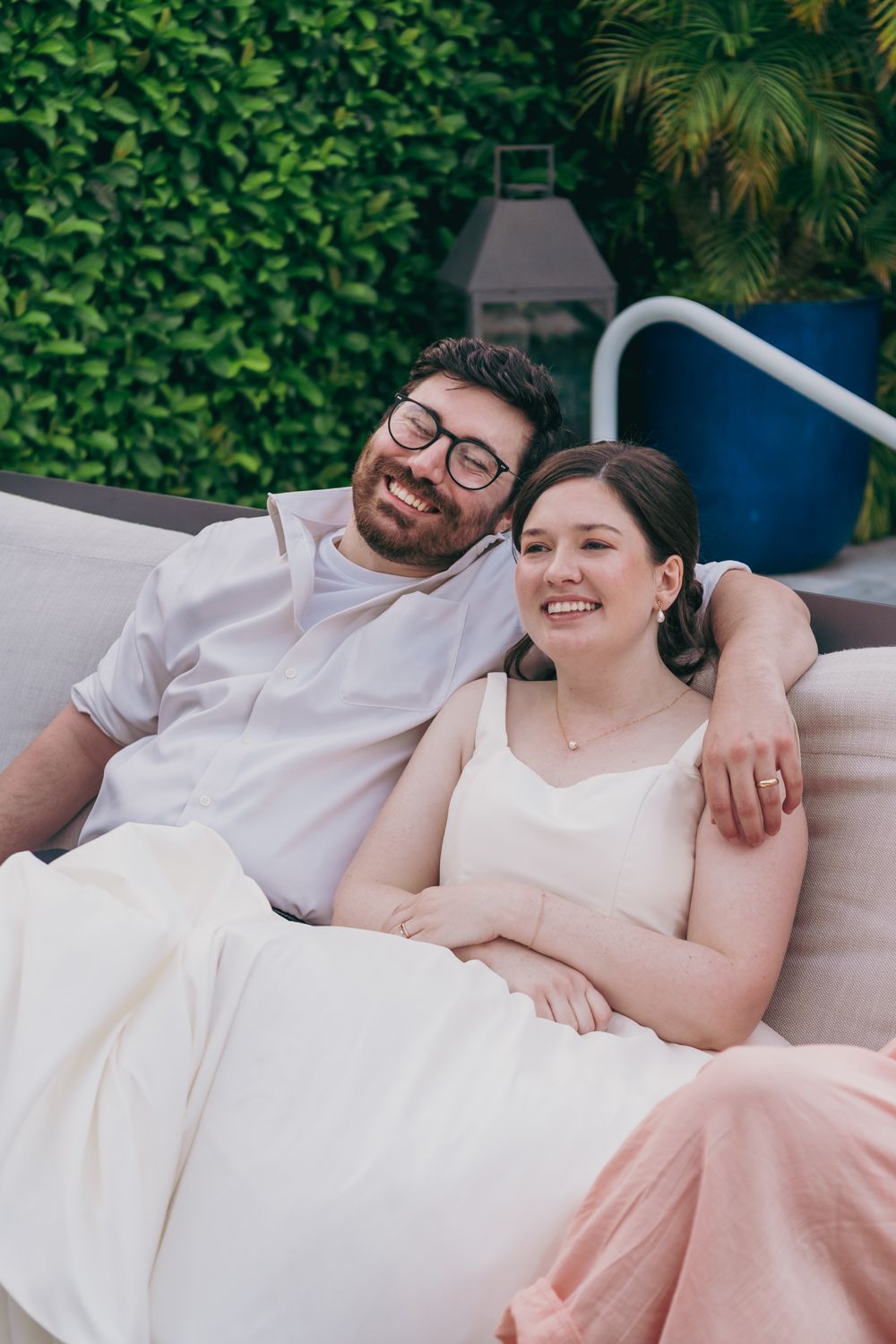 Newlyweds relax and smile together on an outdoor sofa surrounded by green foliage at their wedding.