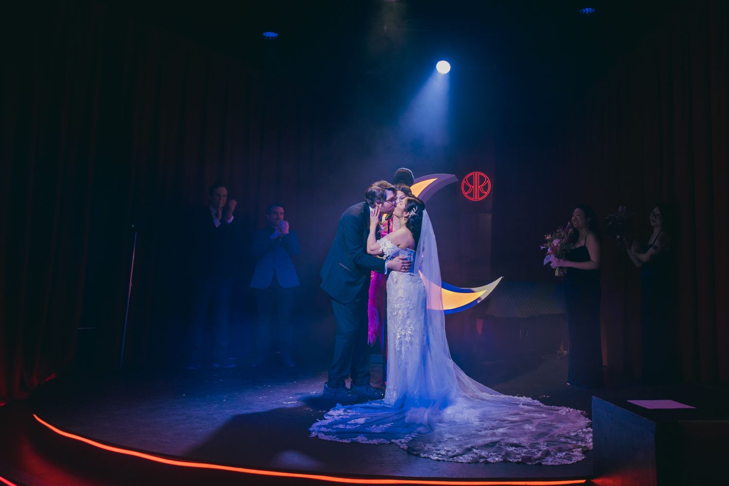 Bride and groom share romantic first dance on stage under blue spotlight with crescent moon backdrop at wedding reception.
