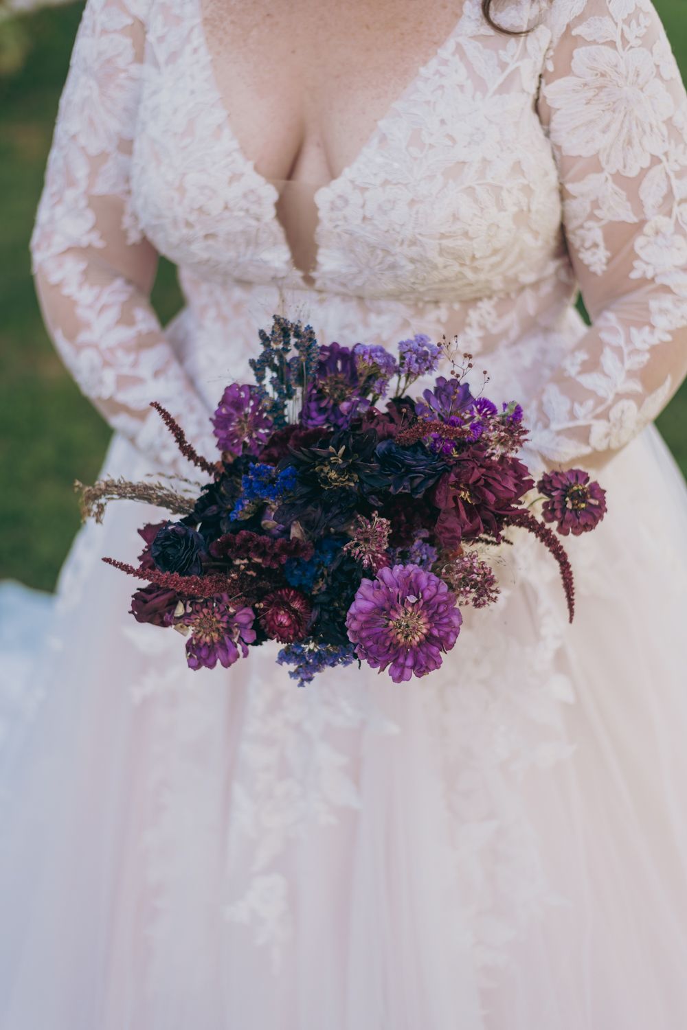 Close-up of bride holding a rich purple and burgundy wedding bouquet against lace gown.