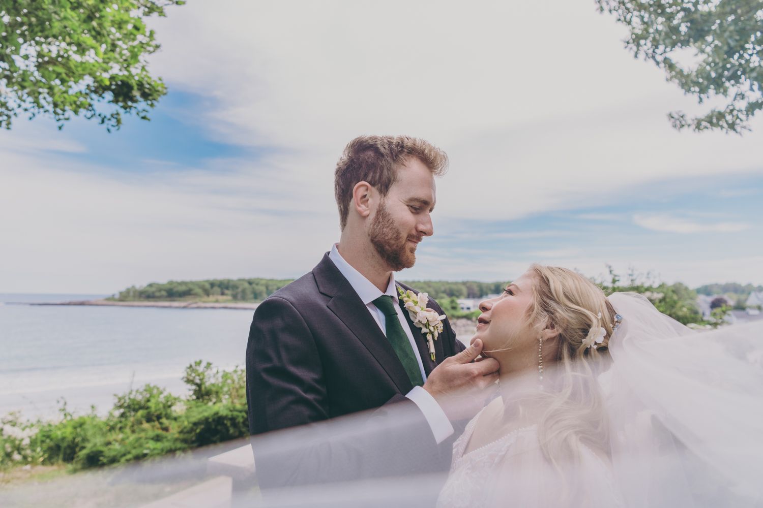 Bride and groom share a romantic moment with veil flowing in the wind by a scenic coastal waterfront.