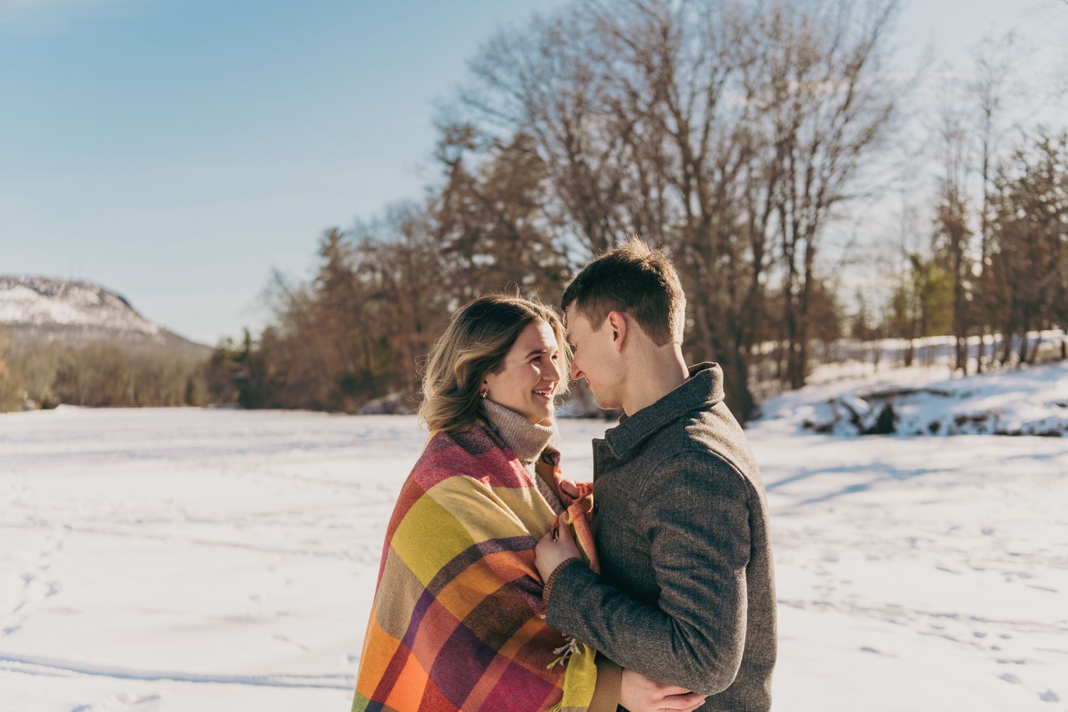Romantic couple wrapped in a colorful plaid blanket sharing a tender moment in a sunny snowy field.