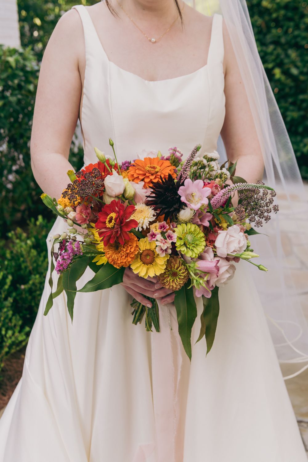 Bride in white gown holds a vibrant wedding bouquet of colorful zinnias, sunflowers, and mixed wildflowers.