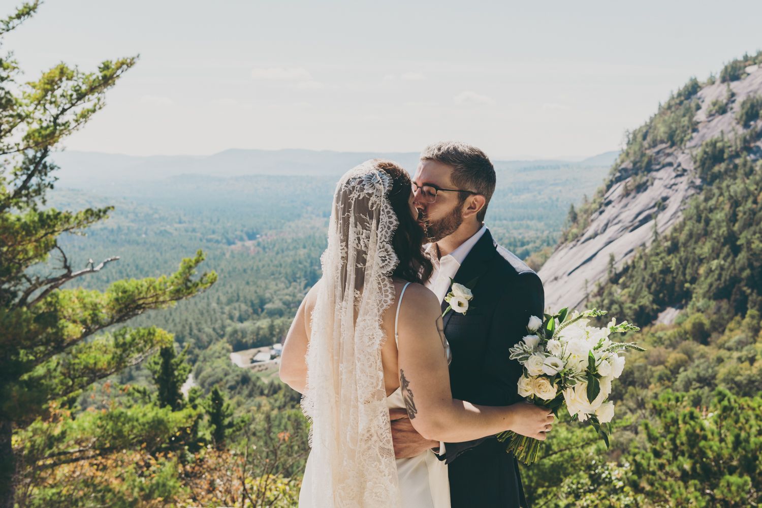 Bride and groom sharing a tender kiss on rocky mountain summit with lush valley views below.