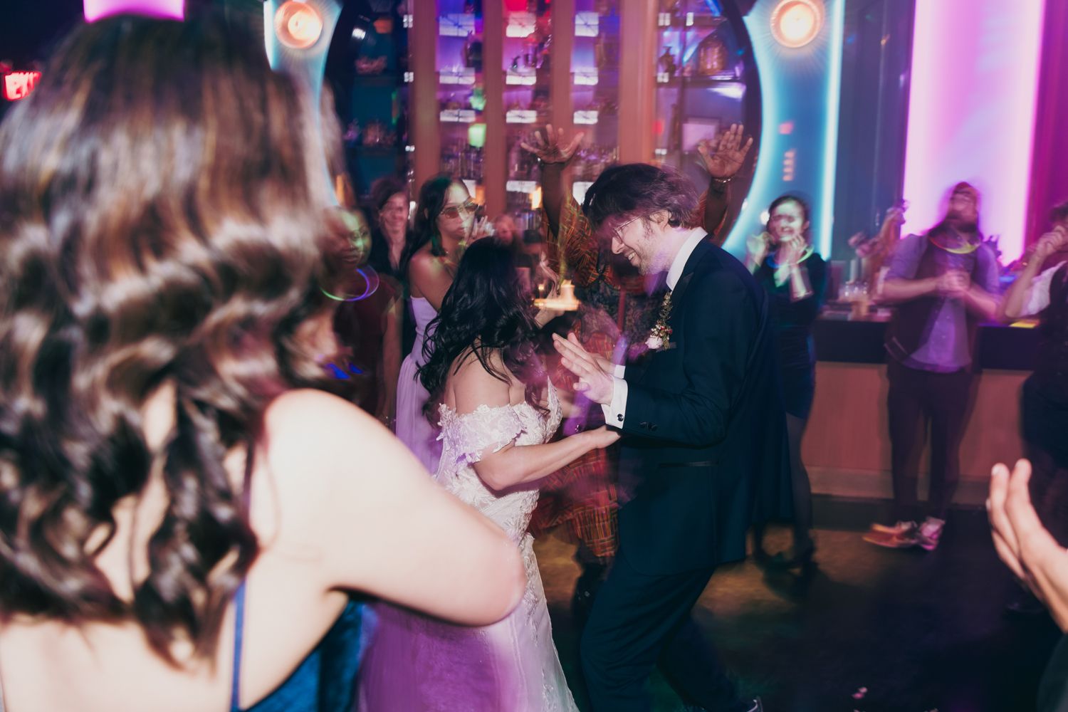 Bride and groom dance joyfully surrounded by celebrating guests on colorful lit reception dance floor.
