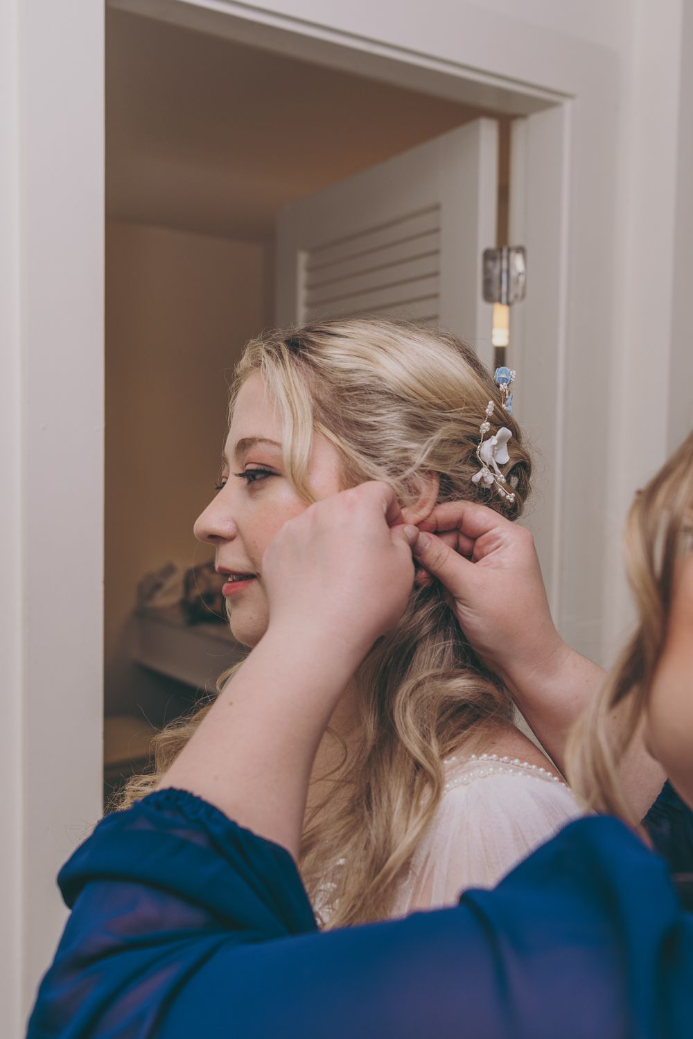 Bridesmaid in blue robe helping blonde bride with floral hair accessory and earring before wedding ceremony.