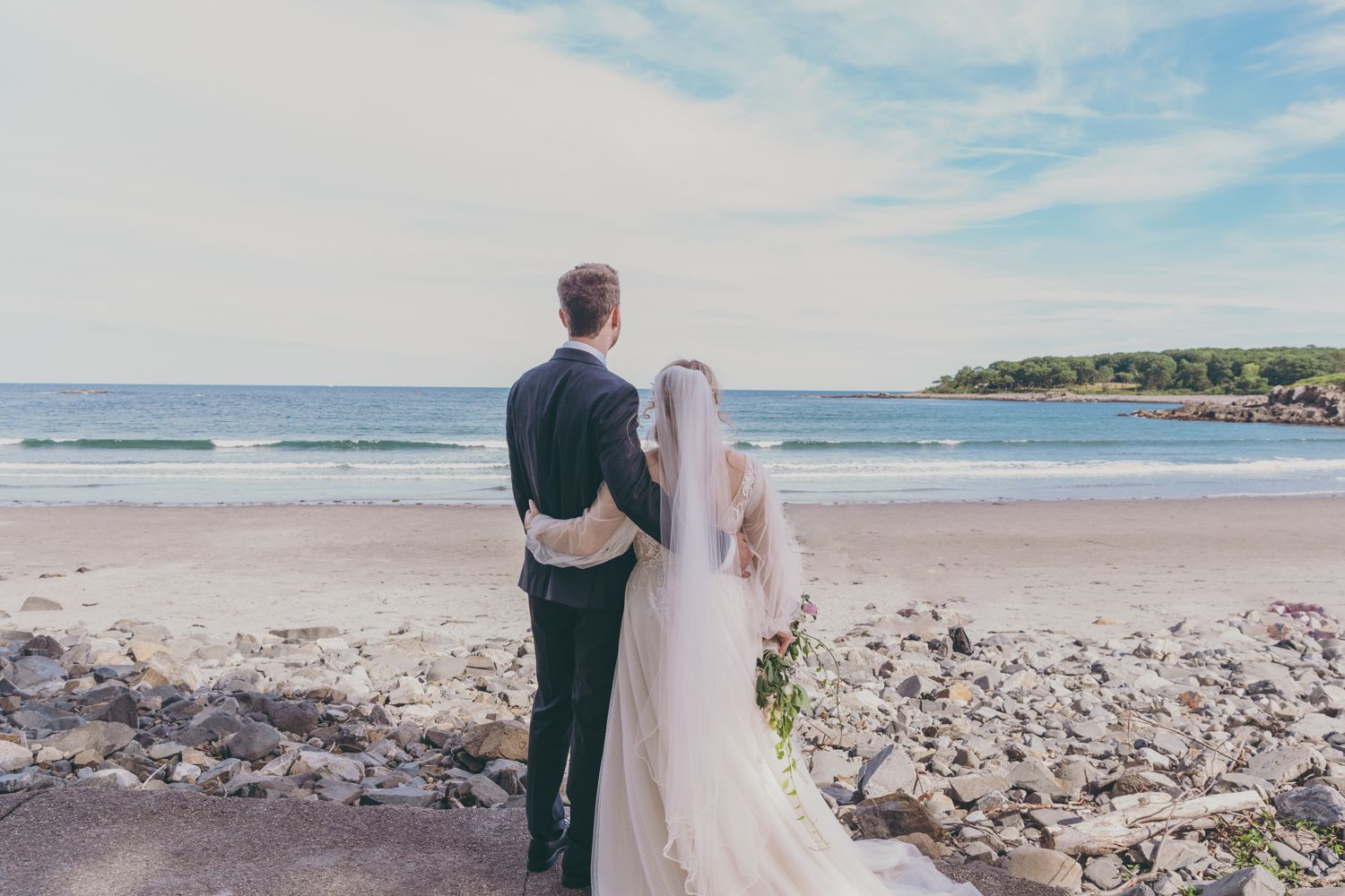 Bride and groom stand hand in hand on a rocky beach, gazing at the ocean on their wedding day.