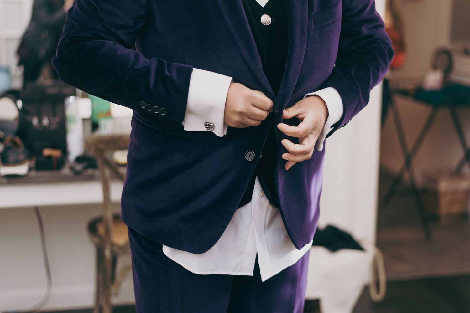 Groom in purple velvet suit fastening buttons on white dress shirt with silver cufflinks.