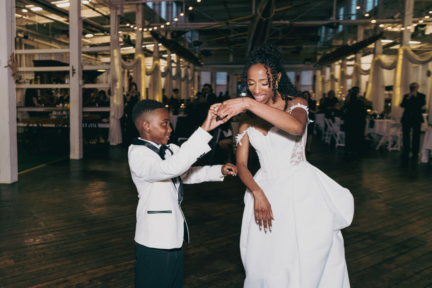 Bride in white wedding gown dancing joyfully with young boy in matching white tuxedo at reception.