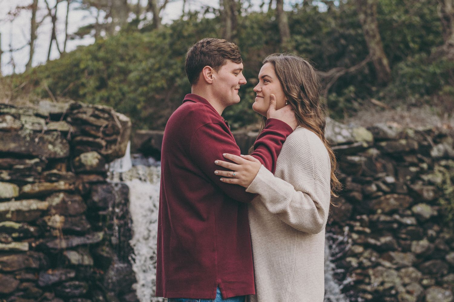 Couple embracing and smiling at each other near a stone wall and waterfall in a romantic outdoor setting.