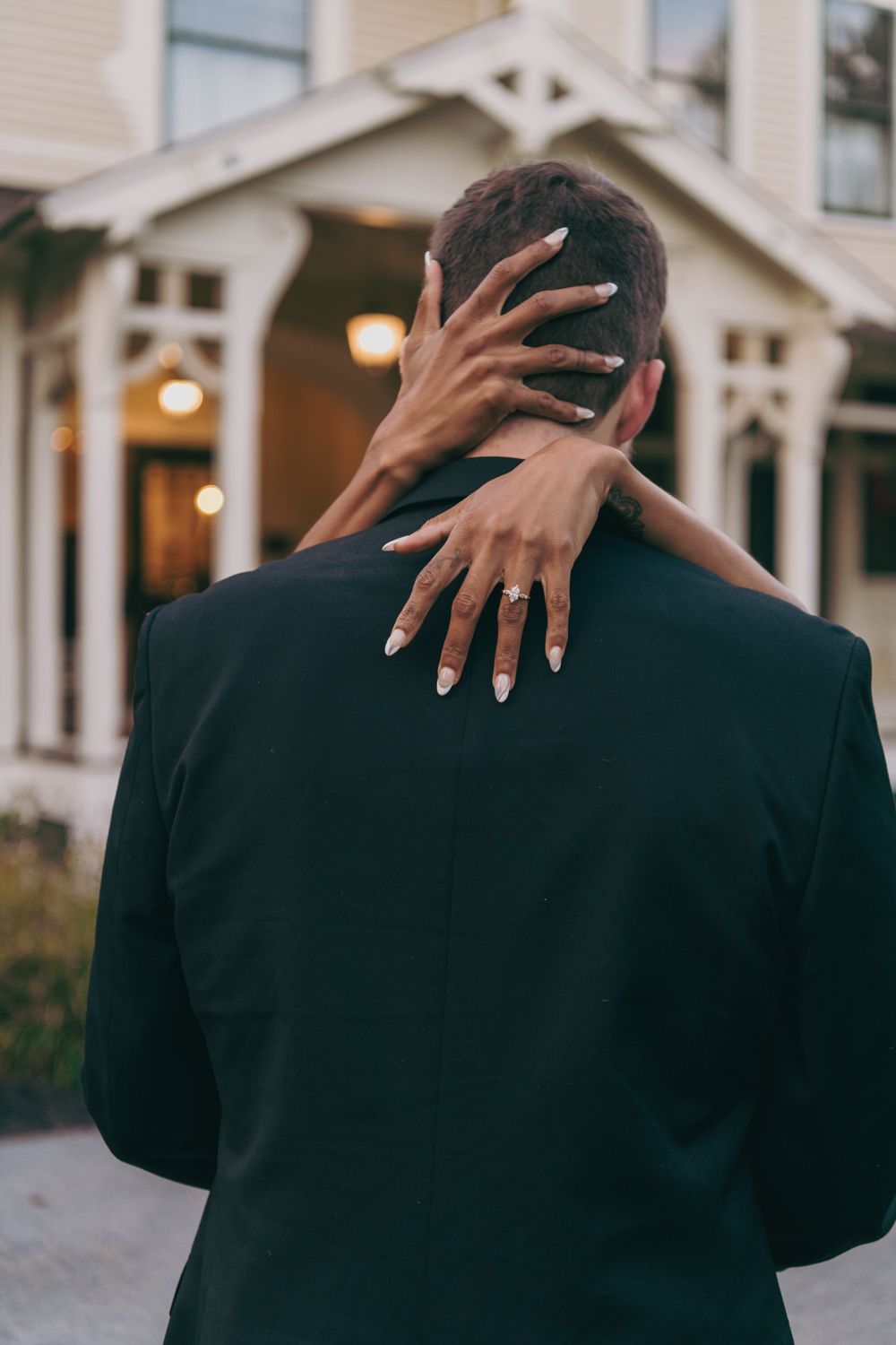 Woman with manicured nails and engagement ring embracing man in black suit from behind near Victorian home.