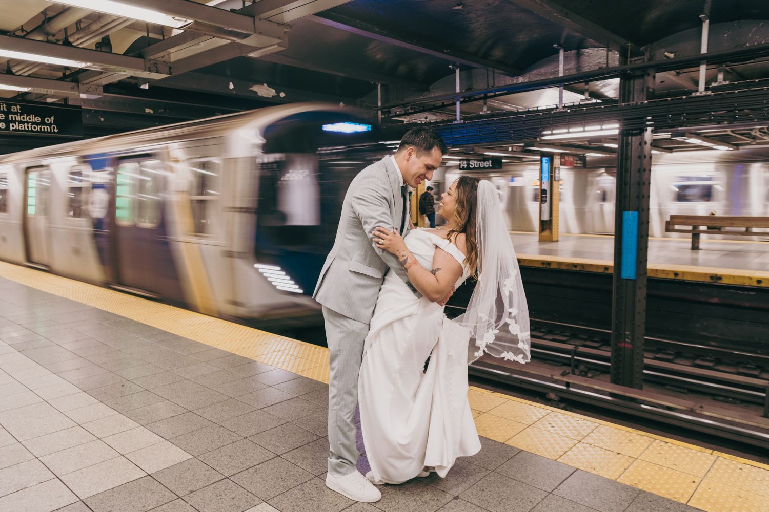 Bride and groom share a romantic dip kiss on a NYC subway platform with a blurred train passing.