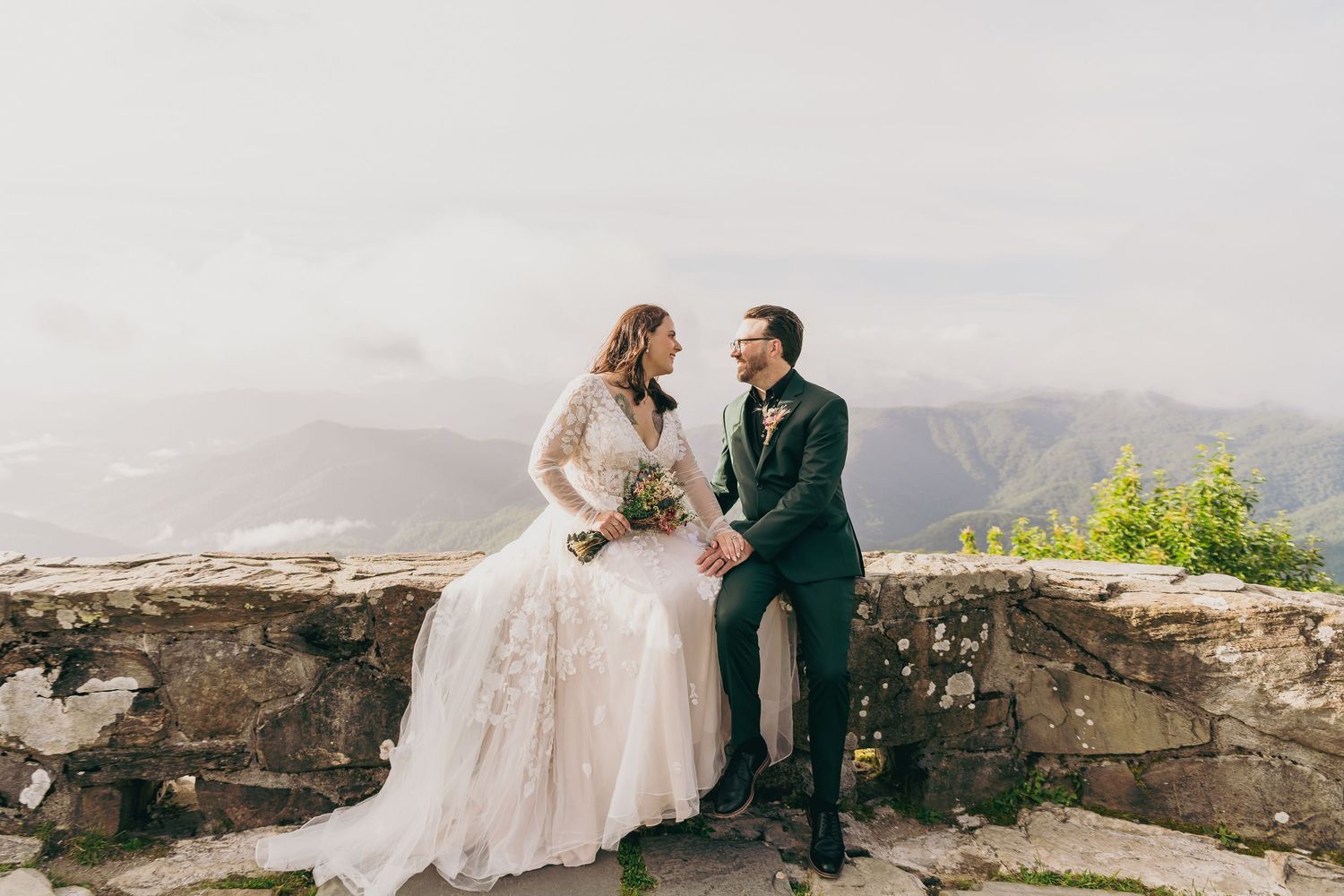 Bride and groom sitting on a stone wall holding hands with misty mountain views on their wedding day.