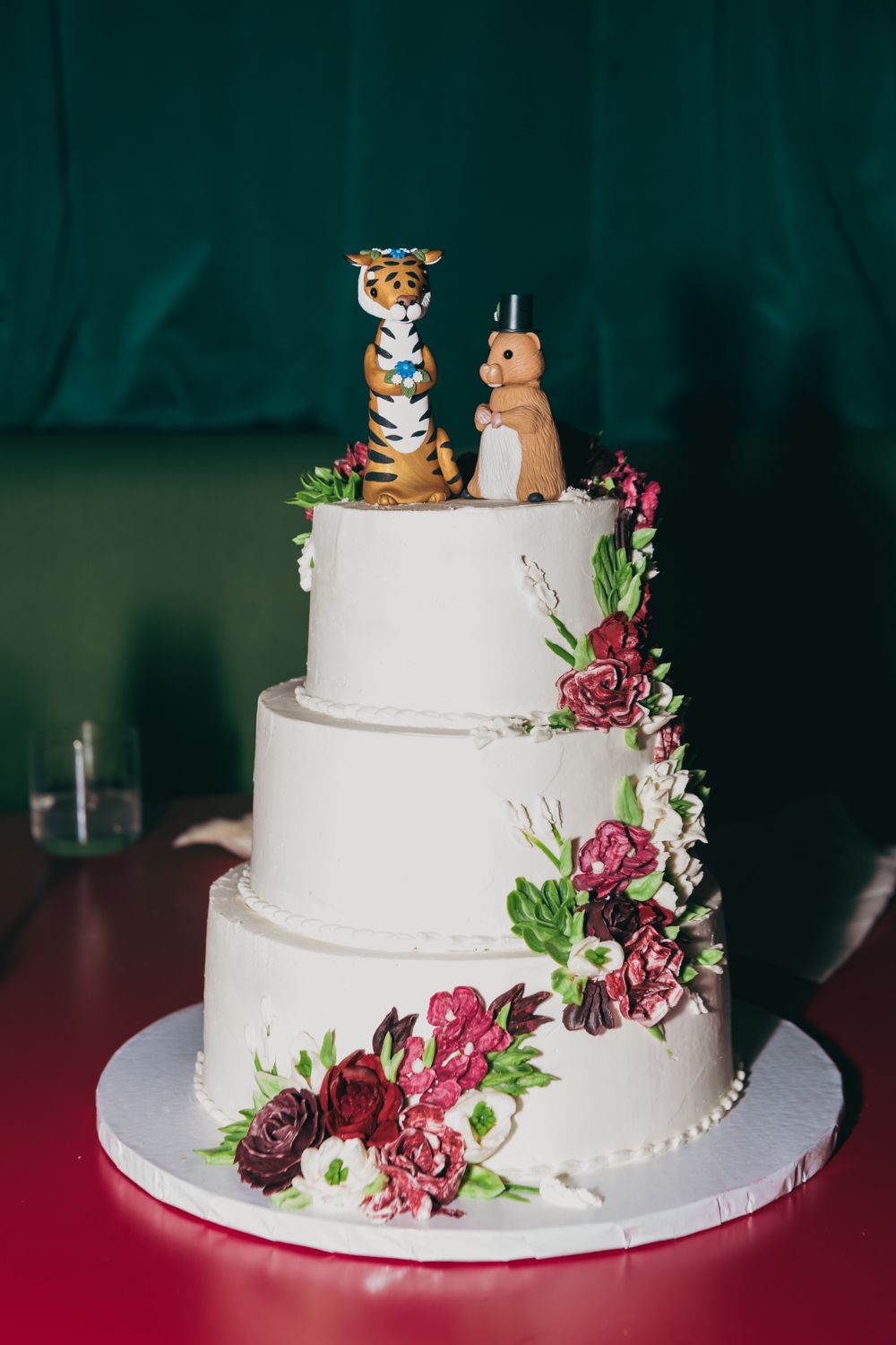 Three-tier white wedding cake with burgundy floral decorations and tiger and monkey animal cake toppers.