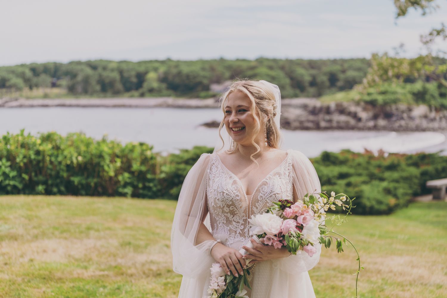 Smiling bride in beaded gown holds pink bouquet outdoors near coastal waterfront on her wedding day.