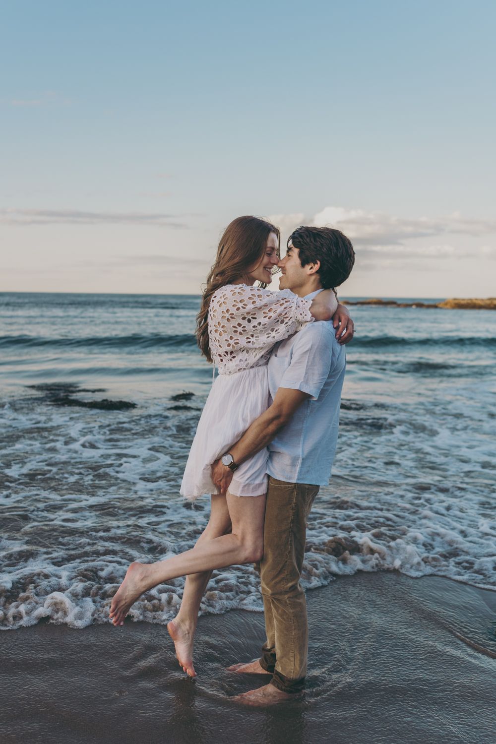 Romantic couple embracing barefoot on a beach at sunset, with ocean waves gently washing around their feet.