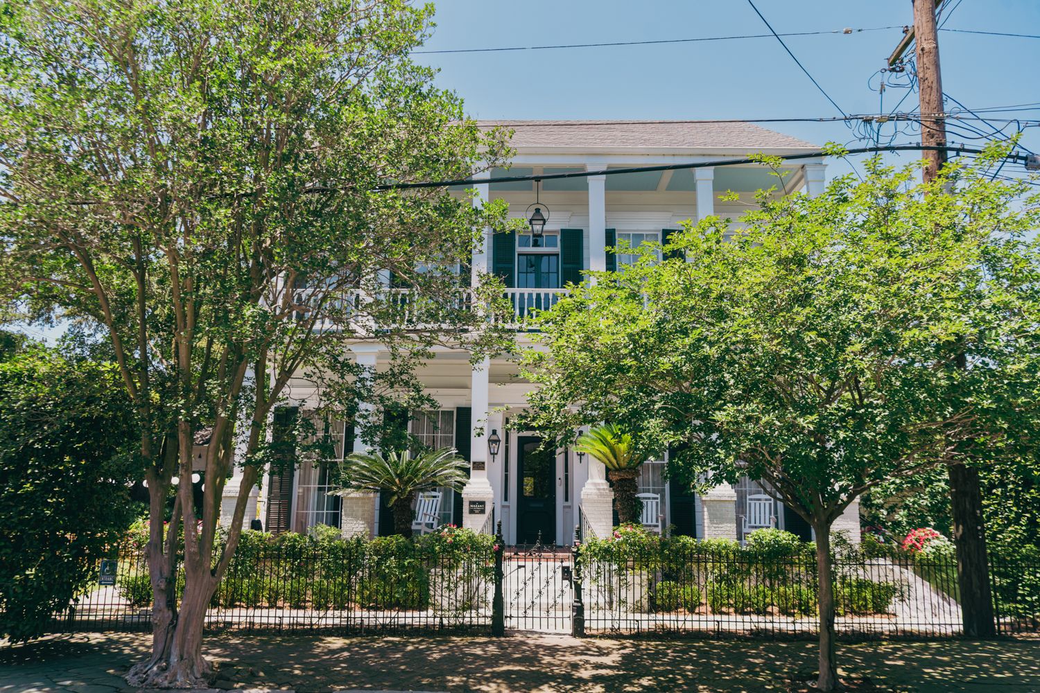 White antebellum mansion with columns, black shutters, and iron fence surrounded by lush green trees.