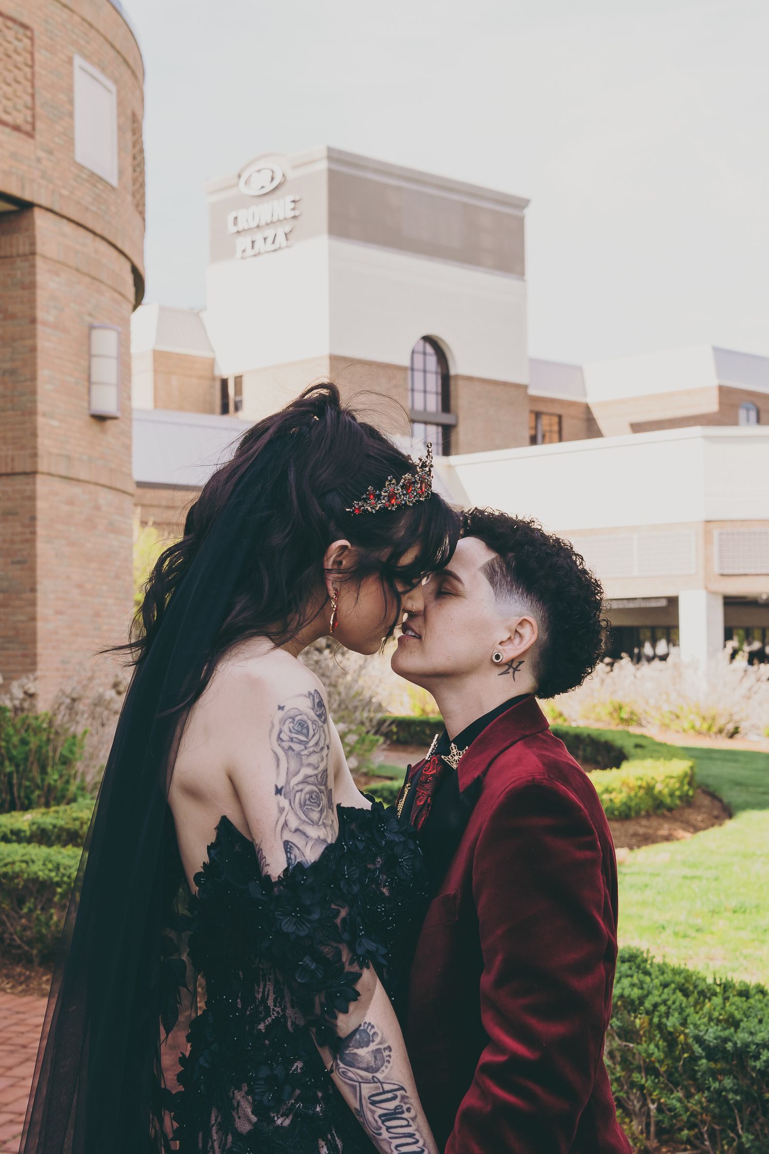 Two people in formal attire sharing a kiss outdoors, one in a black lace gown and one in a burgundy suit.