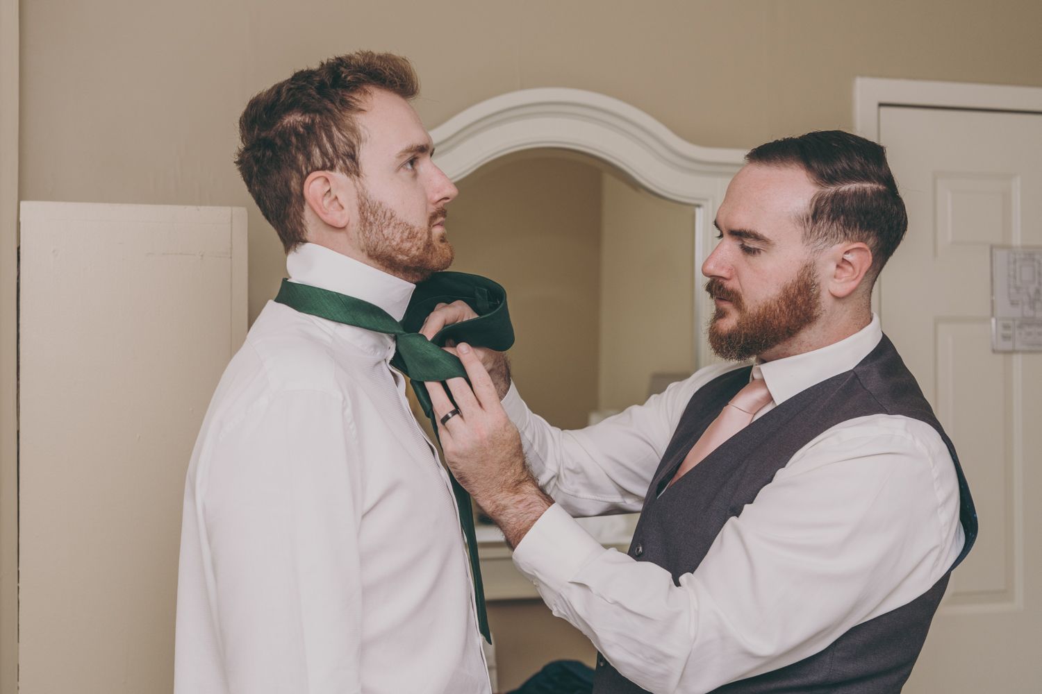 A groomsman adjusts a green bow tie on the groom in a warmly lit room before a wedding ceremony.