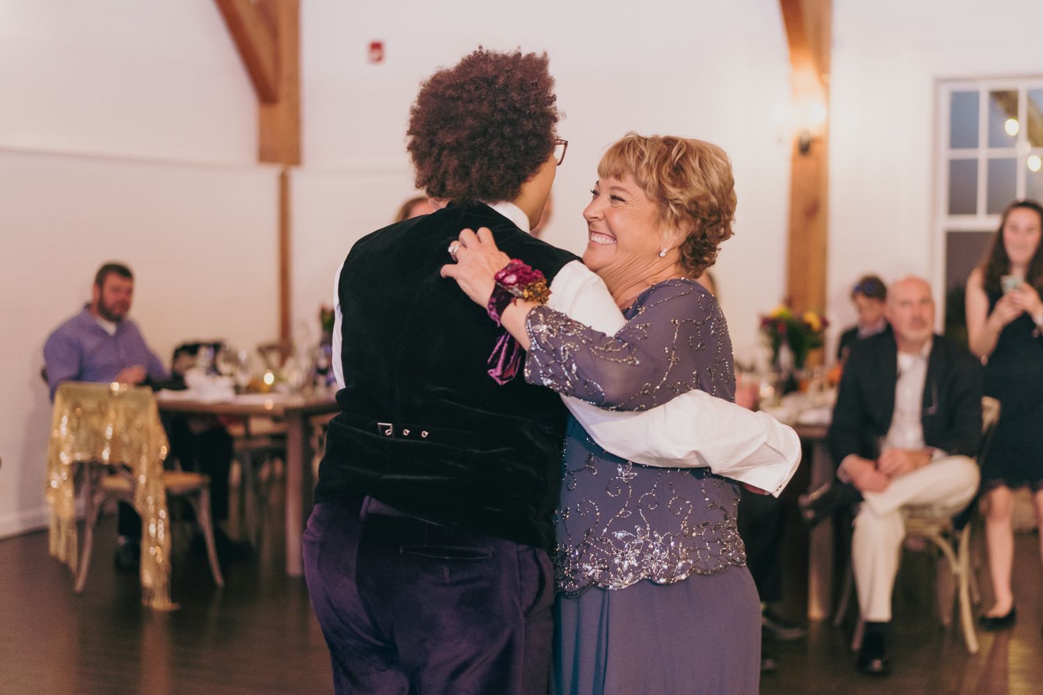 Joyful mother-son dance at wedding reception in bright white venue with smiling guests.