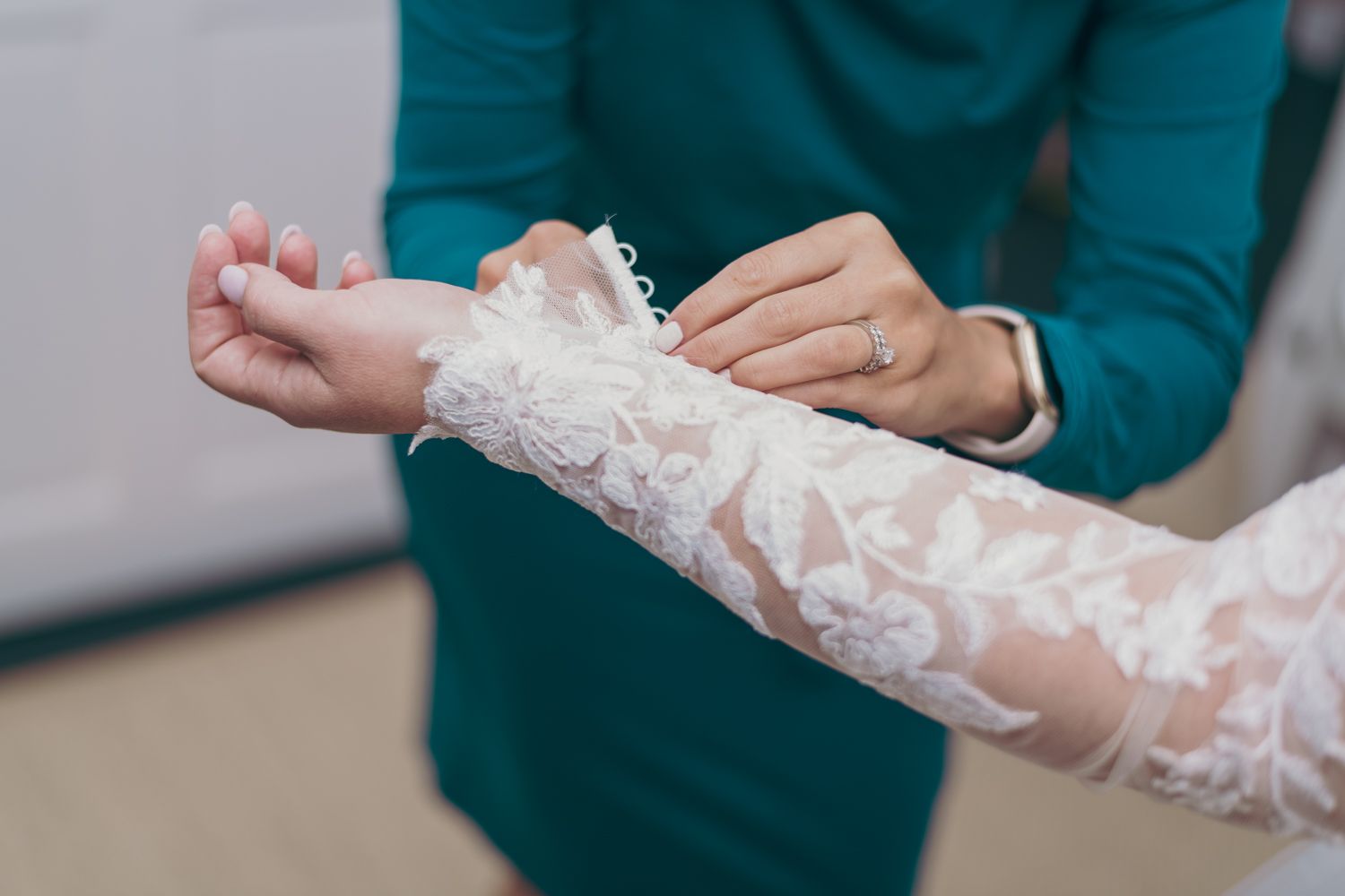 Helper fastening lace wedding dress sleeve button on bride's wrist before ceremony.