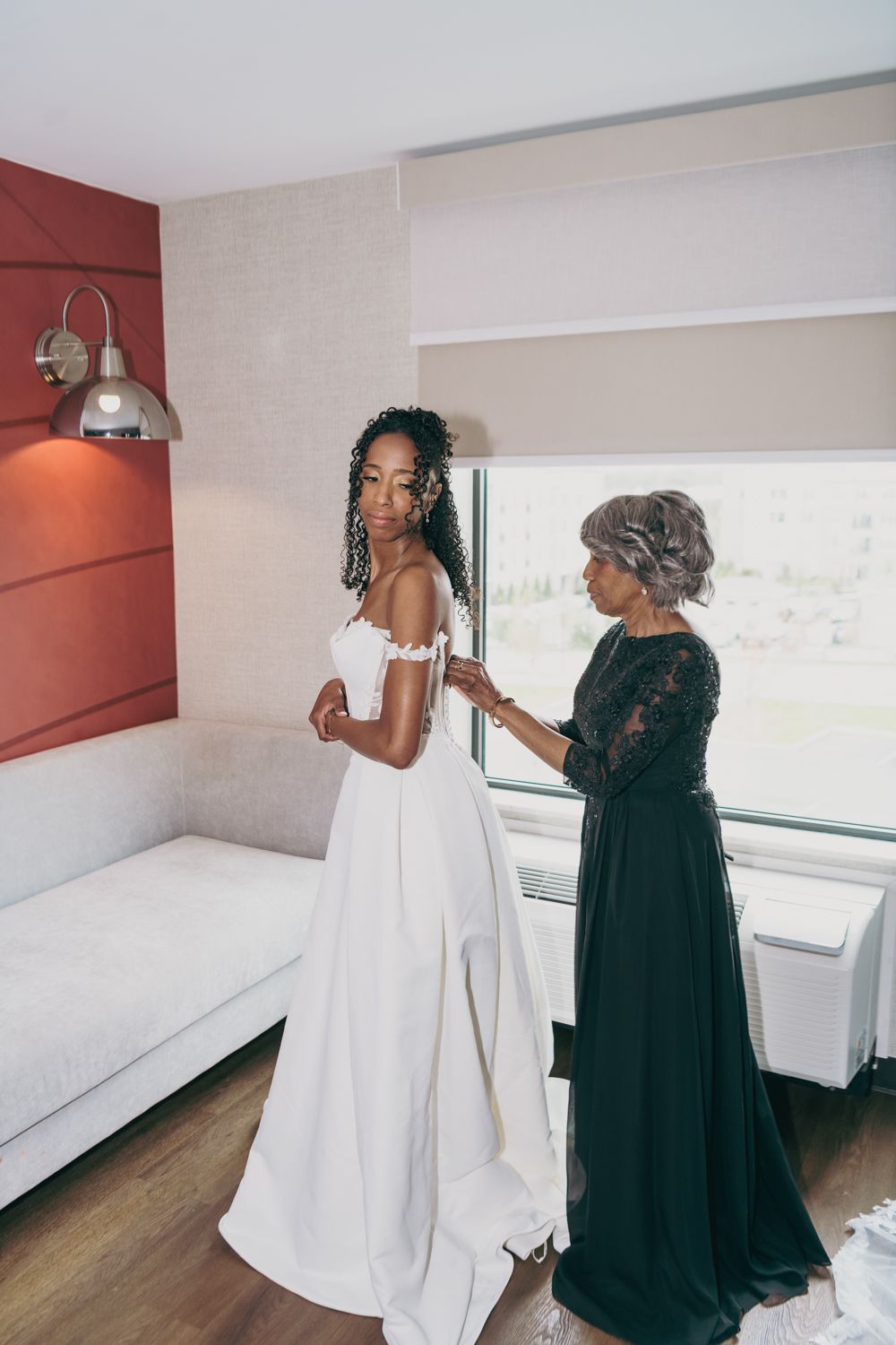 Mother helping bride in white off-shoulder wedding gown get dressed in a bright hotel room.
