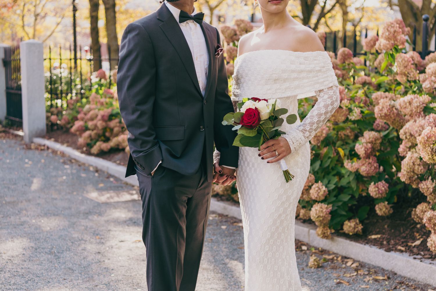 Bride in off-shoulder white gown holding red bouquet standing with groom in navy suit outdoors.