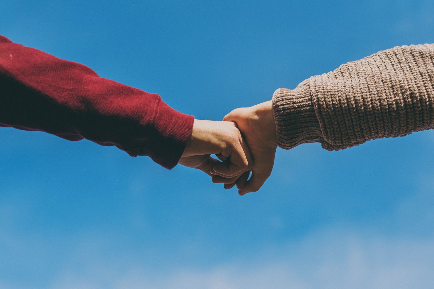 Two people holding hands against a blue sky, one wearing a red sleeve and the other a beige knit sweater.