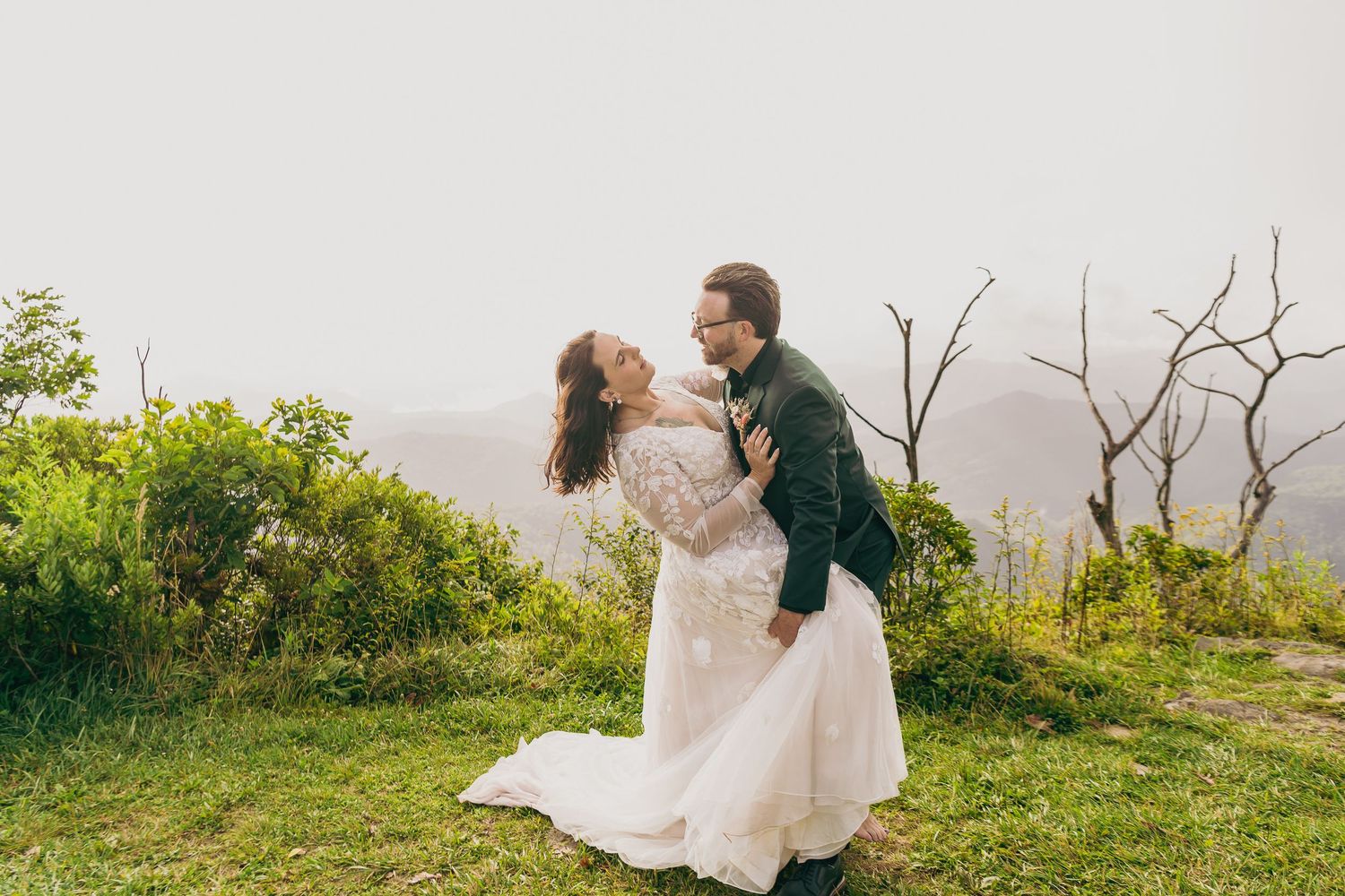 A couple in wedding attire embraces on a misty mountain overlook, surrounded by lush greenery.