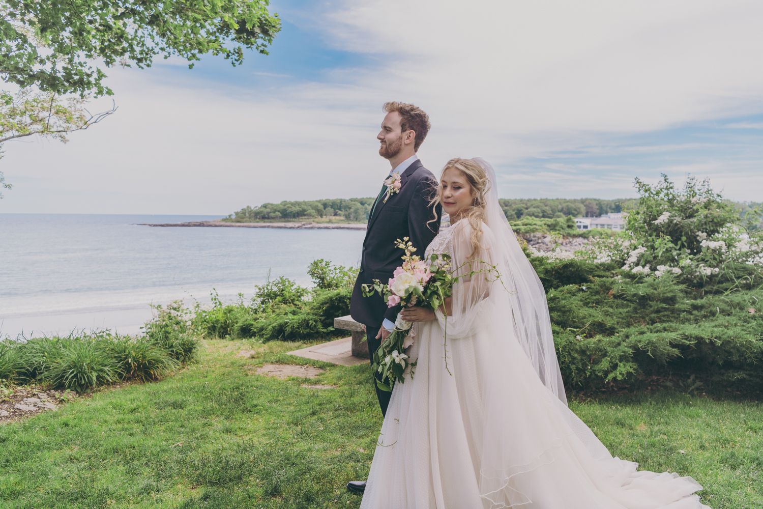 Bride holding floral bouquet and groom pose together overlooking a beautiful ocean landscape on wedding day.