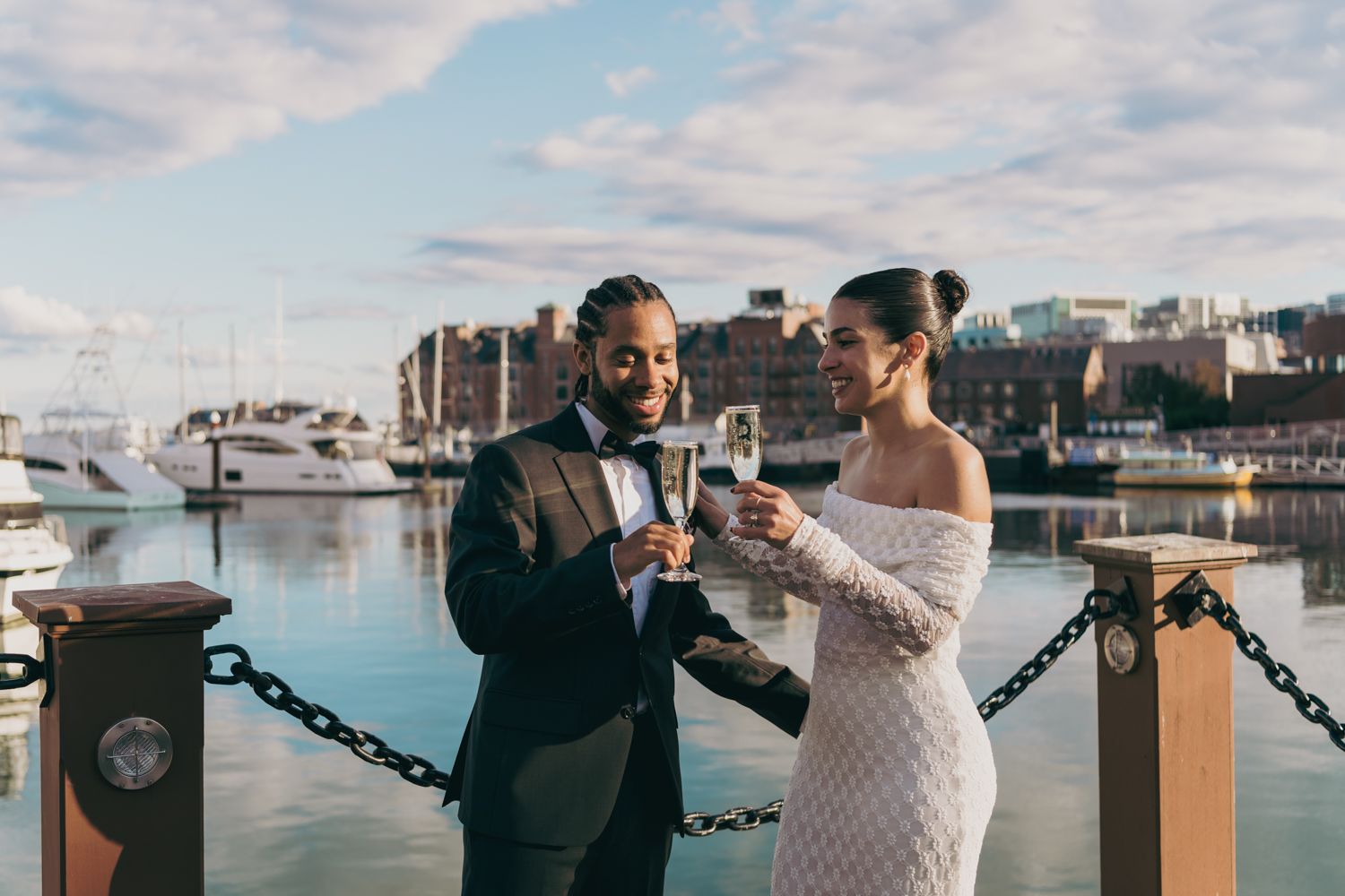 Couple toasting champagne at a waterfront marina, dressed in elegant wedding attire with yachts and city skyline behind them.