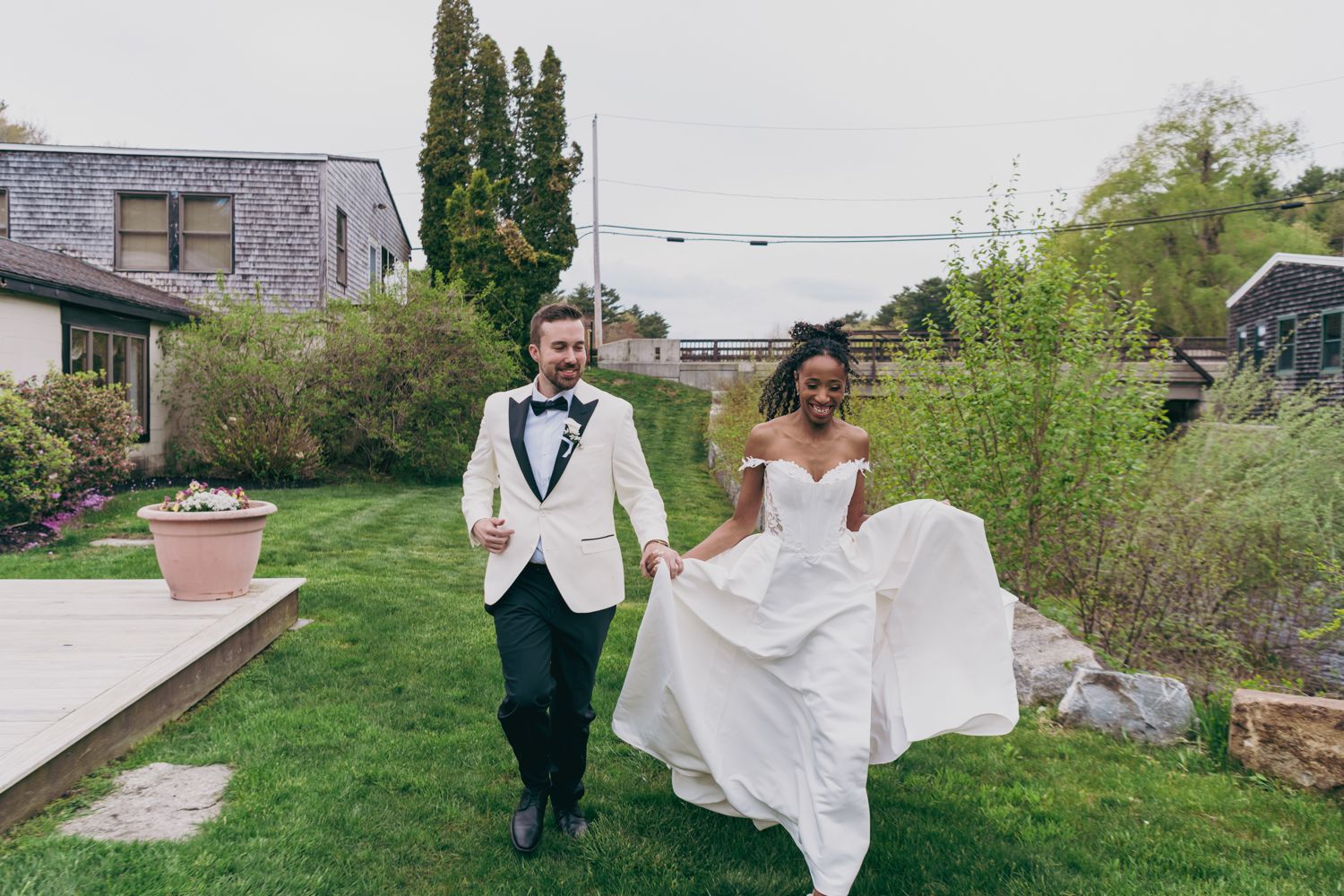 Happy newlywed couple running hand-in-hand across green lawn, bride lifting wedding dress train.