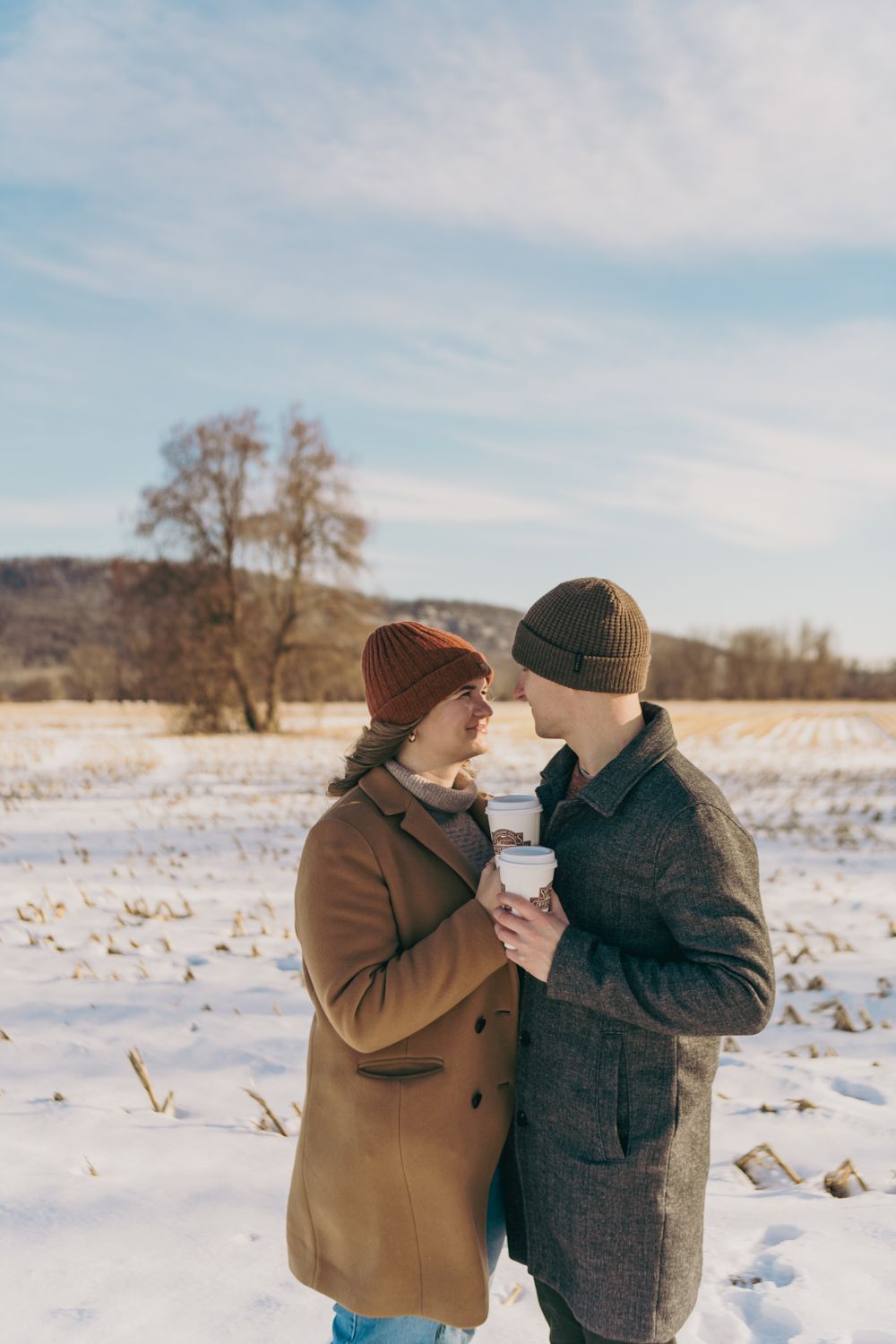 Romantic couple in winter coats and beanies holding coffee cups and almost kissing in a snowy field.
