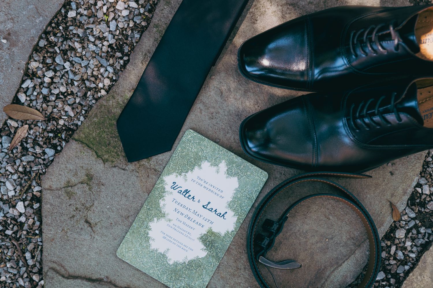 Groom wedding details flat lay with black oxford shoes, dark tie, belt, and elegant Walter Sarah invitation.