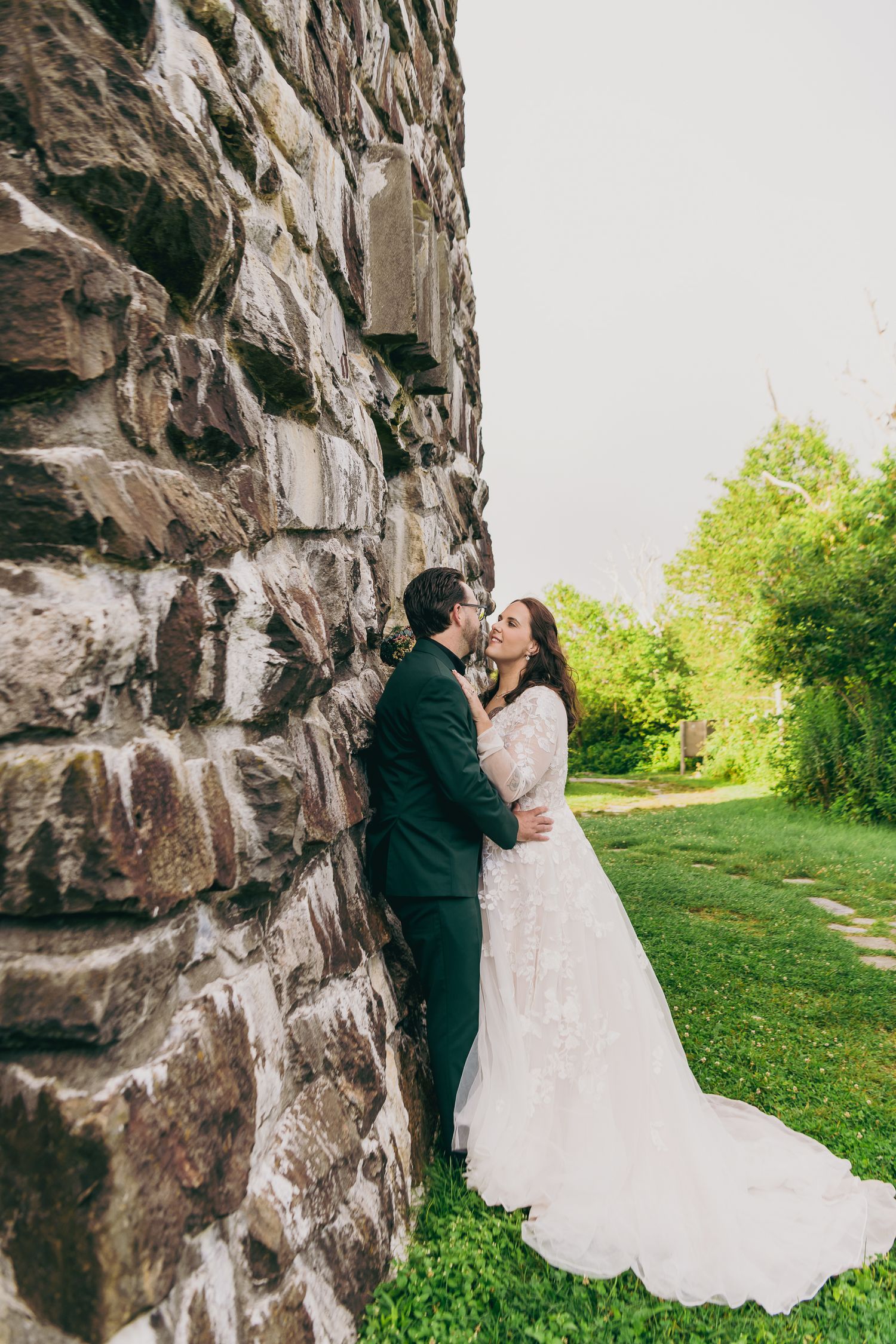 Bride and groom share a romantic kiss against a rustic stone wall surrounded by lush green gardens on their wedding day.